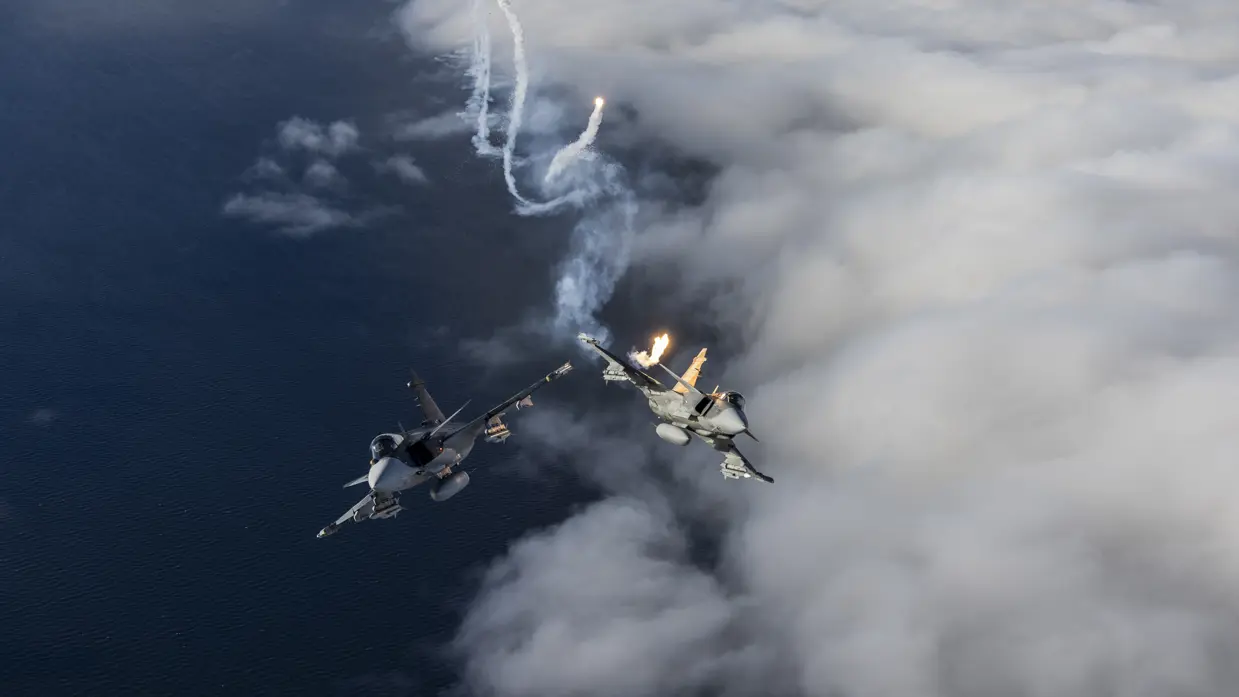 Twee gevechtsvliegtuigen vliegen boven wolken, één vuurt een raket af met rooksporen in de lucht. 