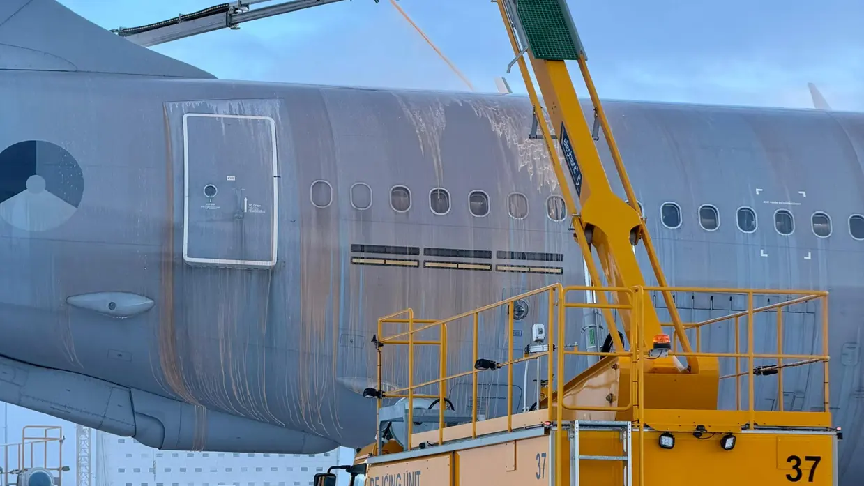 Een gele de-icing truck spuit vanaf hoogte vloeistof op de vleugel van een groot vliegtuig op een besneeuwde luchthaven.