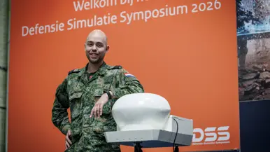 Een militair poseert voor een groot beeldscherm met afbeelding van symposium.