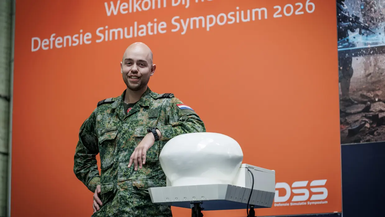 Een militair poseert voor een groot beeldscherm met afbeelding van symposium.