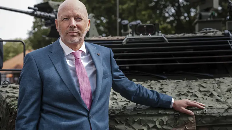 Een kalende man in net pak poseert met zijn hand op een militair voertuig. 