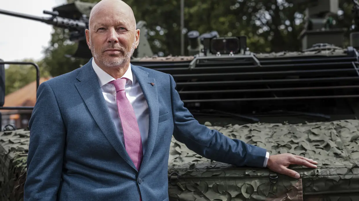 Een kalende man in net pak poseert met zijn hand op een militair voertuig. 