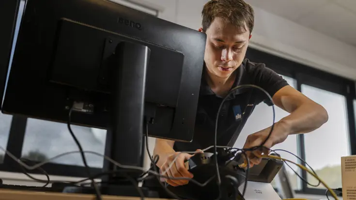 Mick sluit tijdens de opbouw van Falcon Leap beeldschermen en bekabeling aan op een werkplek op Vliegbasis Eindhoven.