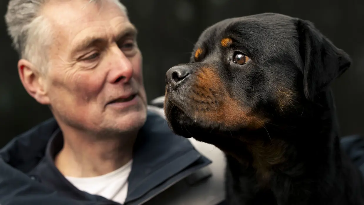 Links een man in uniform. Hij omarmt een grote zwart-bruine hond met opgetrokken oren. Achtergrond toont een bosrijke omgeving met een metalen hek. Rechts een glimlachende man in uniform staat naast een zwarte Rottweiler met open bek. Bosrijke omgeving op de achtergrond, met een ontspannen en vriendelijke sfeer. 