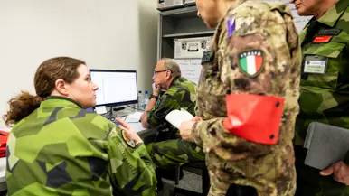 Links: Militairen in uniform werken in een kantoorruimte met laptops en papieren. Een man met Nederlandse vlag op zijn arm staat te luisteren. Kaarten hangen aan de muur. Rechts: Militairen in verschillende uniformen bespreken informatie achter een computer. Een vrouw met Italiaanse vlag op haar mouw kijkt toe, terwijl een man nadenkt.