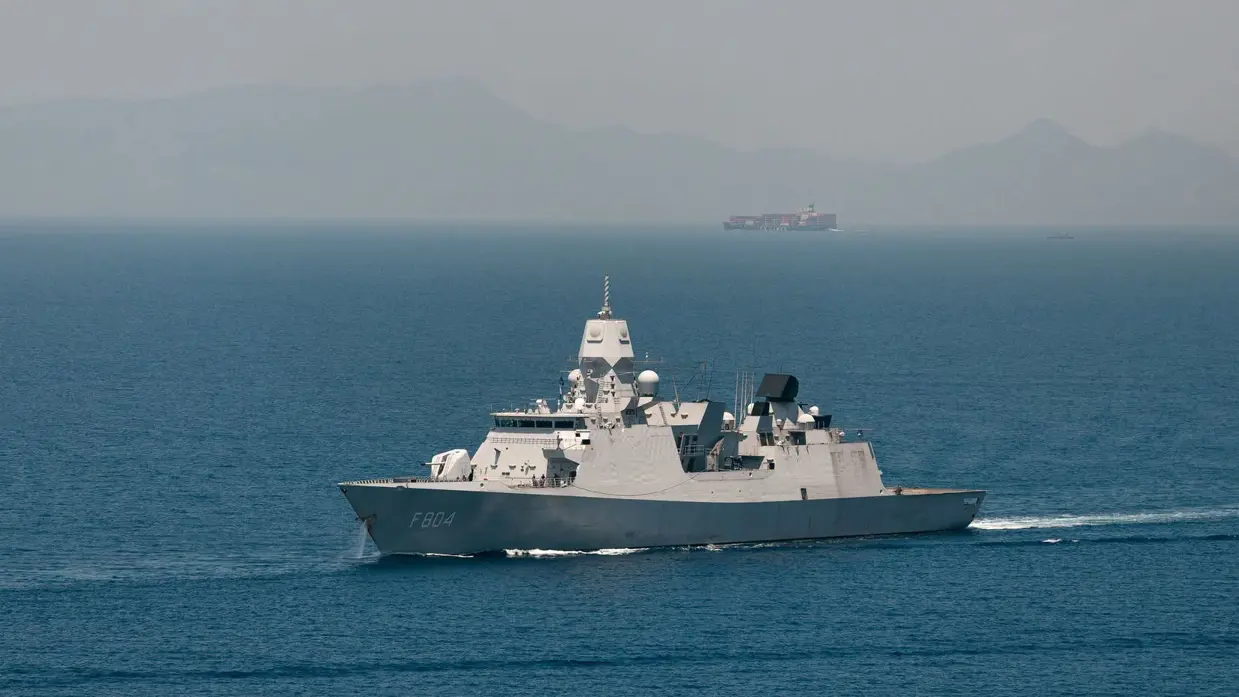 Een marineschip vaart op zee met in de verte een ander schip en hoge bergen.