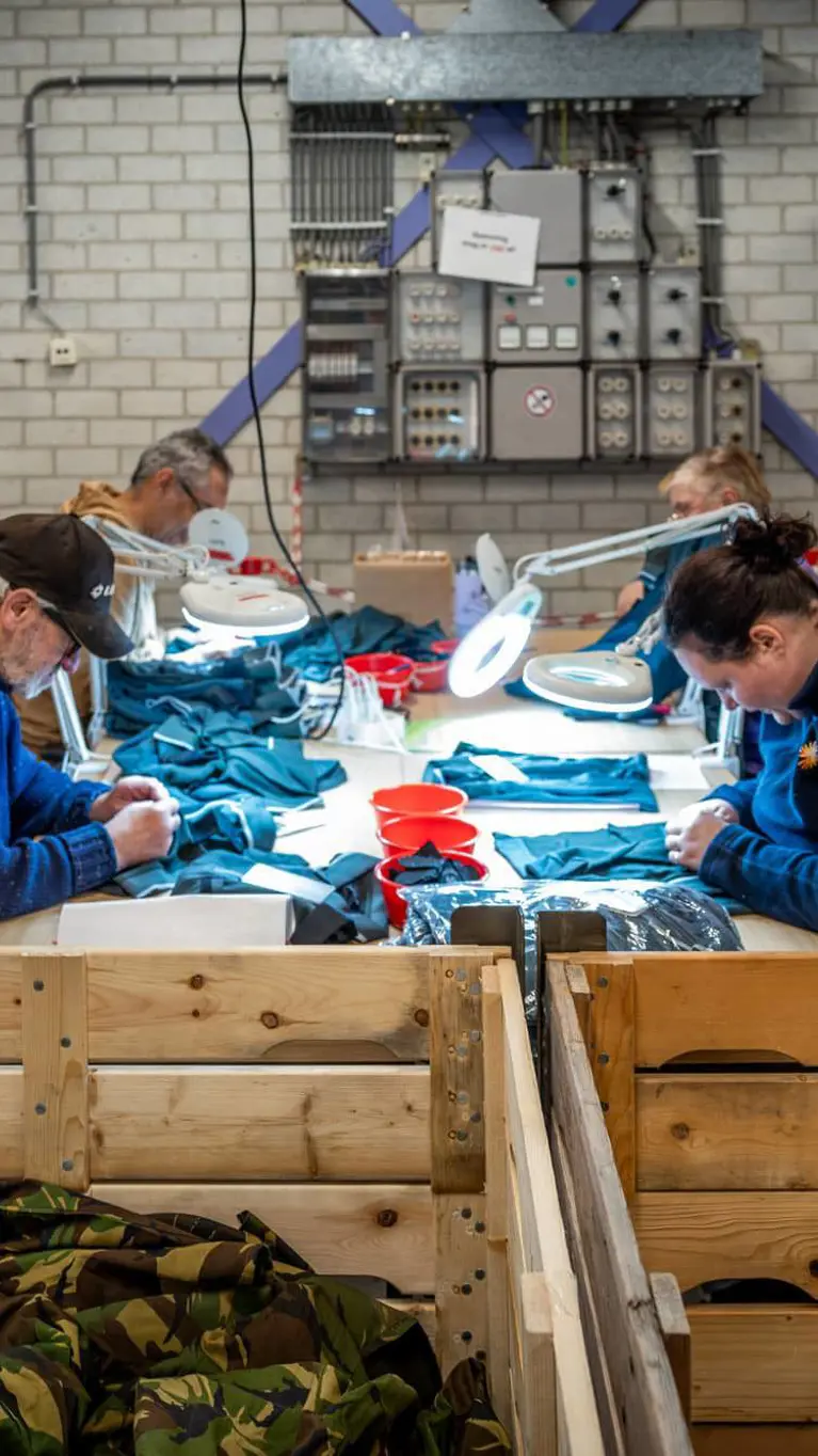 4 personen, van wie een met pet, werken met kleding aan een tafel.  