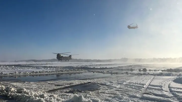 Twee helikopters vliegen laag over een besneeuwde, modderige en ijsbedekte landingsbaan onder een heldere, blauwe hemel met zonnig licht. 
