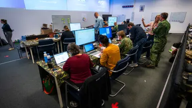 Drie burgermedewerkers en militairen zitten in een grote ruimte achter beeldschermen met aan de wand enkele heel grote beeldschermen.