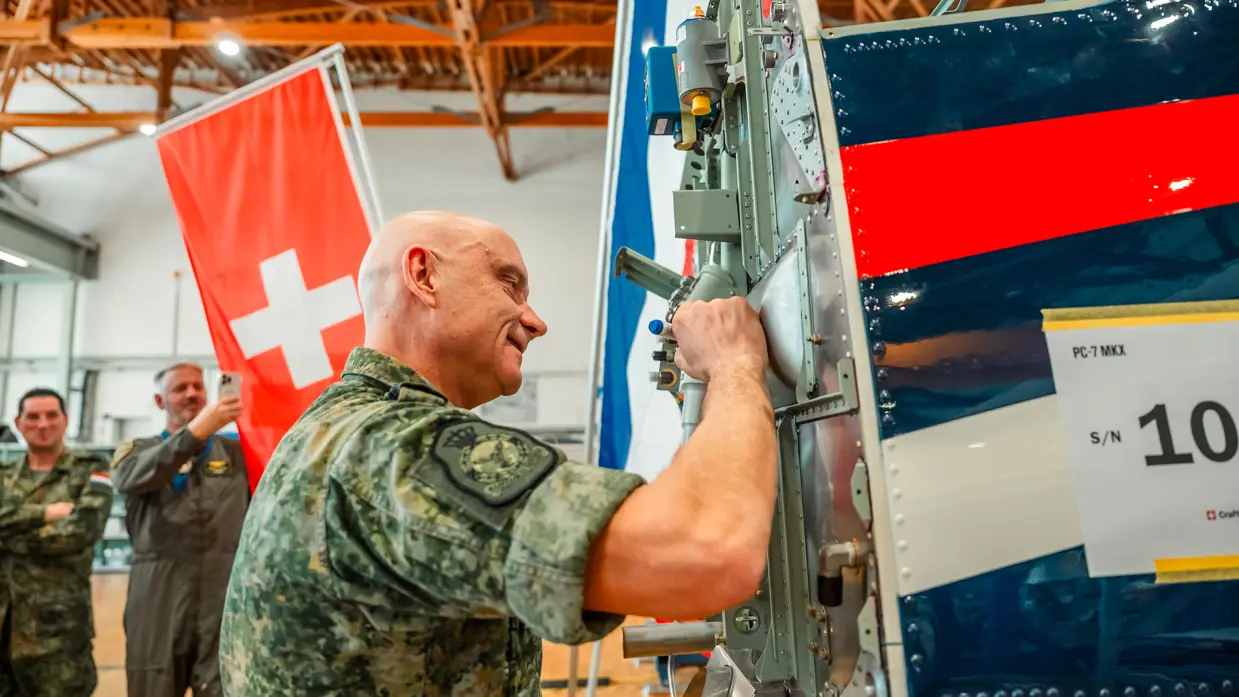 Een militair zet zijn handtekening op de romp van een lesvliegtuig, bijgestaan door andere genodigden.