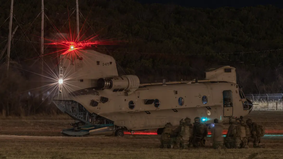 In duisternis groepje knielende militairen naast een Chinook met openstaande achterklep.