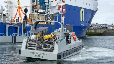 Links een grijs bootje met achterop verschillende drones. Dit vaart langs een groot vrachtschip. Daarnaast een gele, raketvormige drone met voorop een witte bol. Deze is ook te zien achterop het varende bootje.