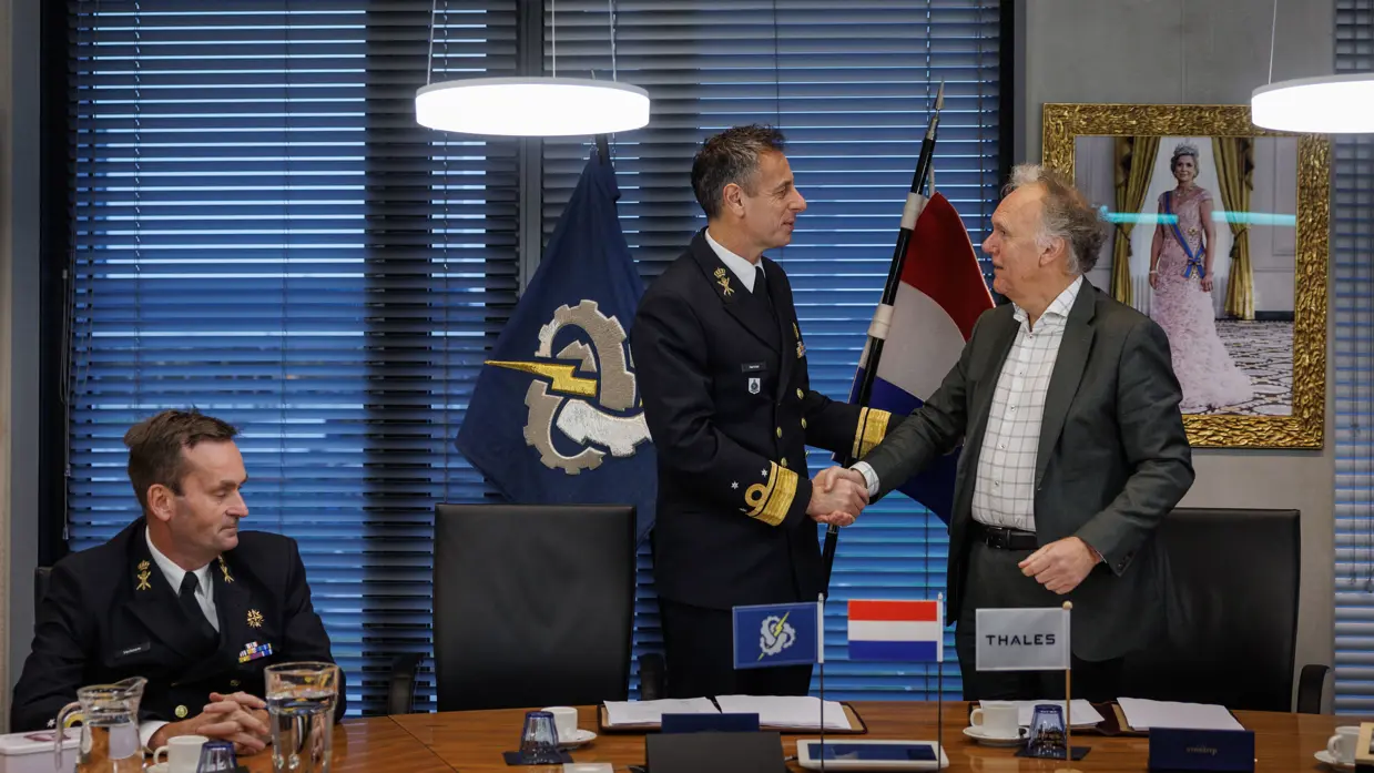 Twee mannen schudden elkaar de hand, van wie één in militair tenue. Ze staan achter een tafel. Op de achtergrond hangt een schilderij van de koningin en staan vlaggen, die ook in het klein op het bureau staan.