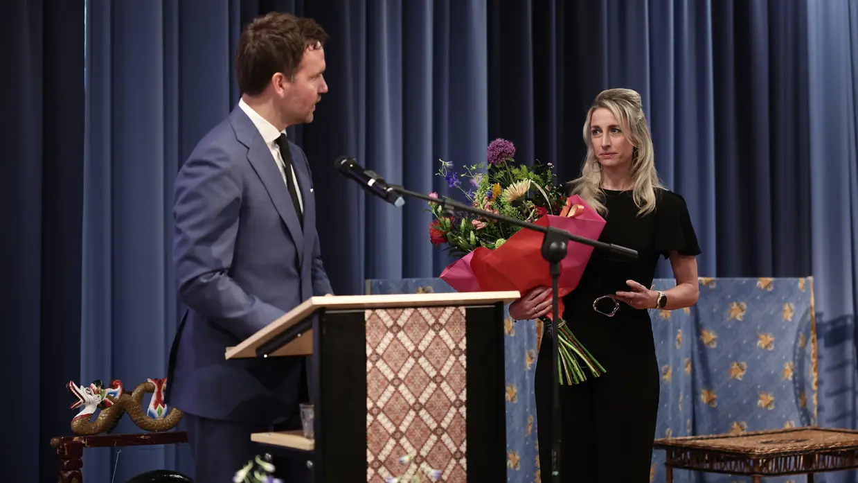 De staatssecretaris, in donkerblauw pak, staat achter een microfoon en spreekt Amber toe. Zij staat ernaast en luistert naar wat hij te zeggen heeft. In haar hand een bos bloemen.