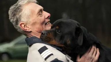 Links een man in uniform. Hij omarmt een grote zwart-bruine hond met opgetrokken oren. Achtergrond toont een bosrijke omgeving met een metalen hek. Rechts een glimlachende man in uniform staat naast een zwarte Rottweiler met open bek. Bosrijke omgeving op de achtergrond, met een ontspannen en vriendelijke sfeer. 