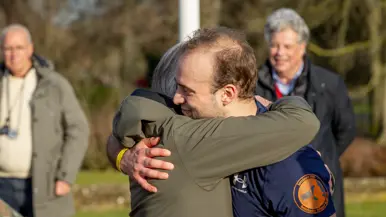 Links een groep mensen in militaire kleding loopt rennend over een keienpad, omringd door bomen en een gebouw. Aan de zijkant houden twee mannen een spandoek vast. Het is een zonnige dag. Rechts: twee mannen omarmen elkaar hartelijk buiten in de zon. De ene draagt een groene jas, de andere een donkerblauw sportshirt met een oranje logo. Op de achtergrond staan twee oudere mannen. 