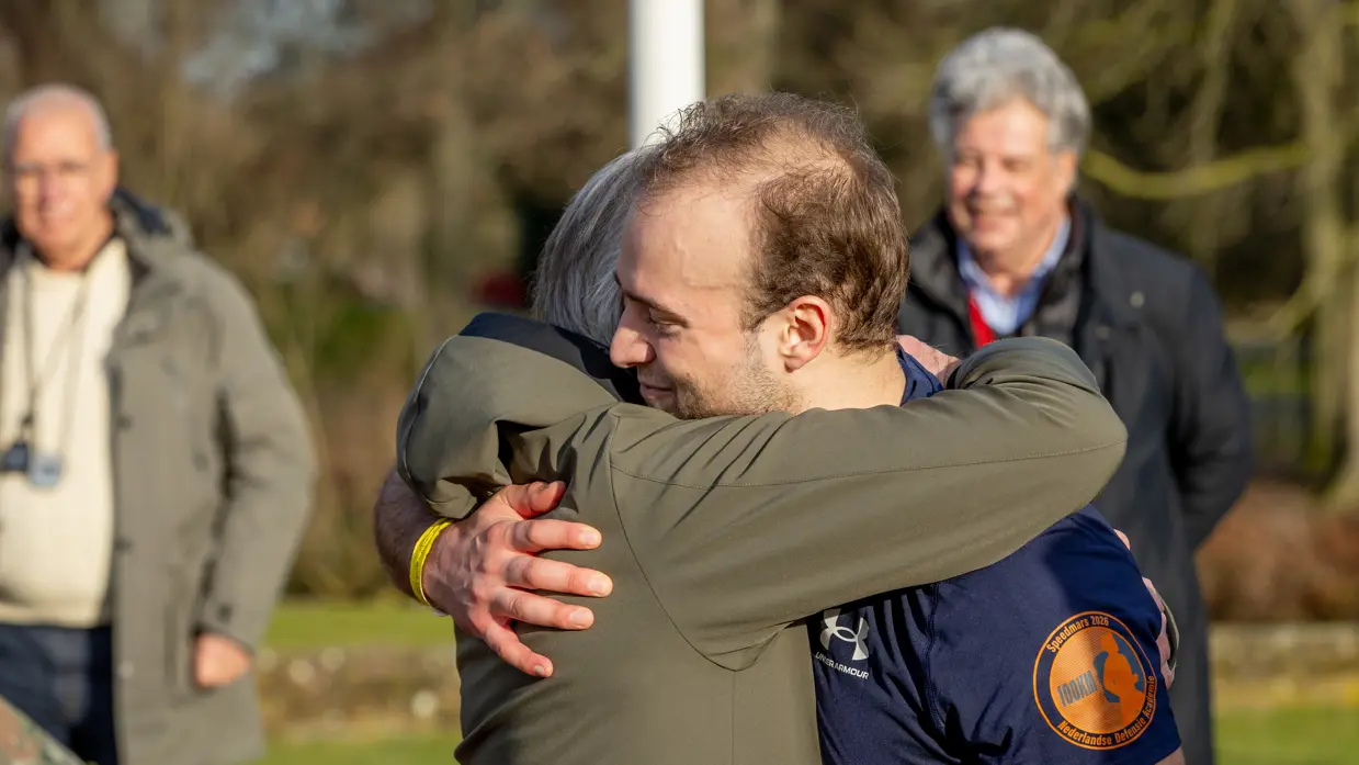 Links een groep mensen in militaire kleding loopt rennend over een keienpad, omringd door bomen en een gebouw. Aan de zijkant houden twee mannen een spandoek vast. Het is een zonnige dag. Rechts: twee mannen omarmen elkaar hartelijk buiten in de zon. De ene draagt een groene jas, de andere een donkerblauw sportshirt met een oranje logo. Op de achtergrond staan twee oudere mannen. 
