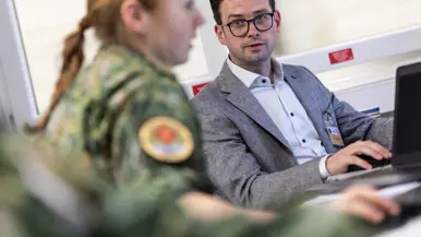 Links: Een man in pak met bril en identiteitskaart werkt op een laptop. Een vrouw in militair uniform met een medisch embleem staat naast hem. Rechts: drie mannen lopen glimlachend door een gang: een in donkerblauw uniform, een in Nederlands militair tenue met medisch embleem, en een in Amerikaans legeruniform. 