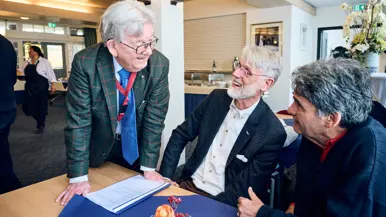 Een man in pak spreekt met twee heren die aan een tafel zitten in een zaal.