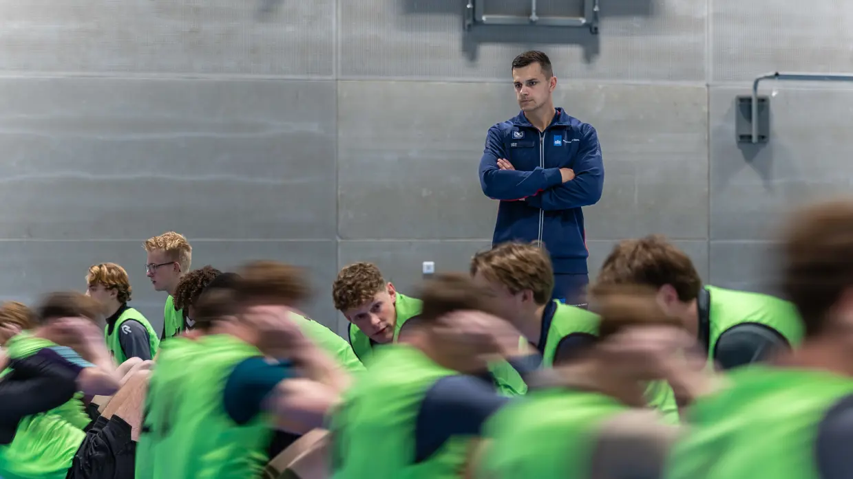 Jongeren in gele hesjes voeren diverse sportonderdelen uit in een sportzaal. 
