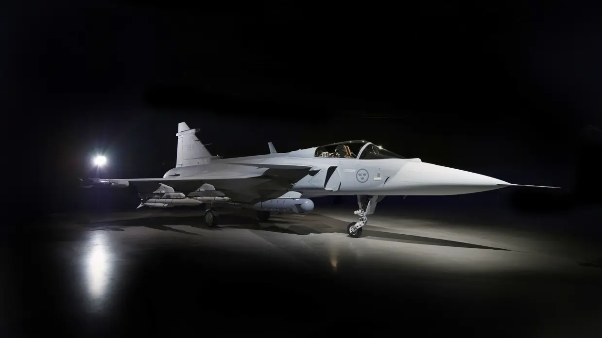 Een straaljager in een donkere hangar, verlicht door een felle lamp. Het vliegtuig staat stil op de grond, met een slanke, puntige neus en wapens onder de vleugels. 