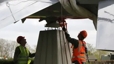 Twee arbeiders in veiligheidskleding bevestigen een groot metalen voorwerp onder de vleugel van een vliegtuig. 
