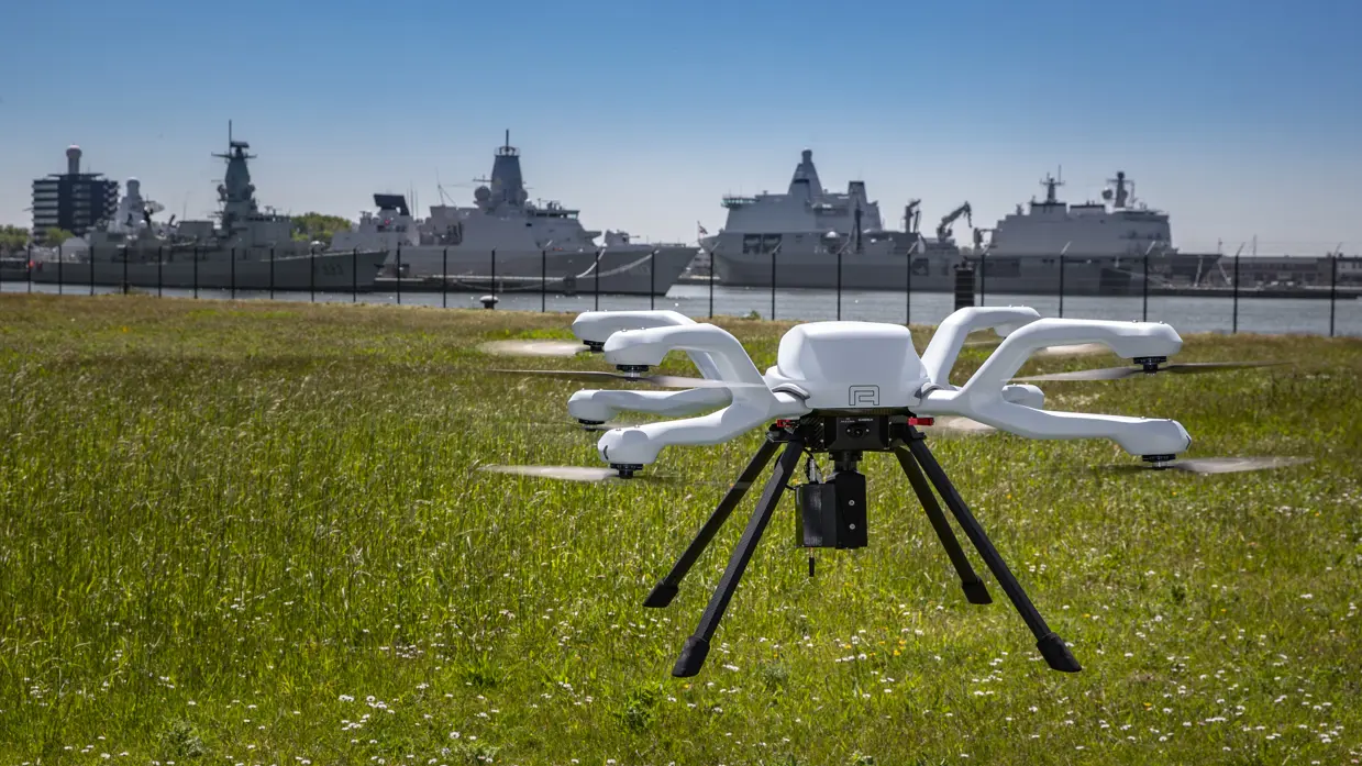 een vliegende drone op de voorgrond, enkele fregatten op de achtergrond
