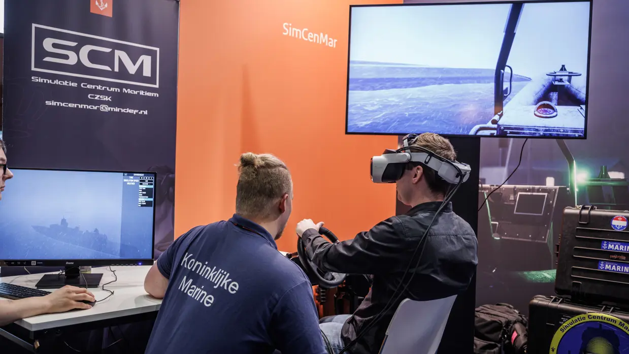 Een persoon in een t-shirt van marine praat in een gameroom tegen iemand in burgerkleding met VR-bril op.