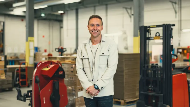 Kevin draagt een witte blouse en kijkt in de camera. Achter hem zie je een grijze loods.