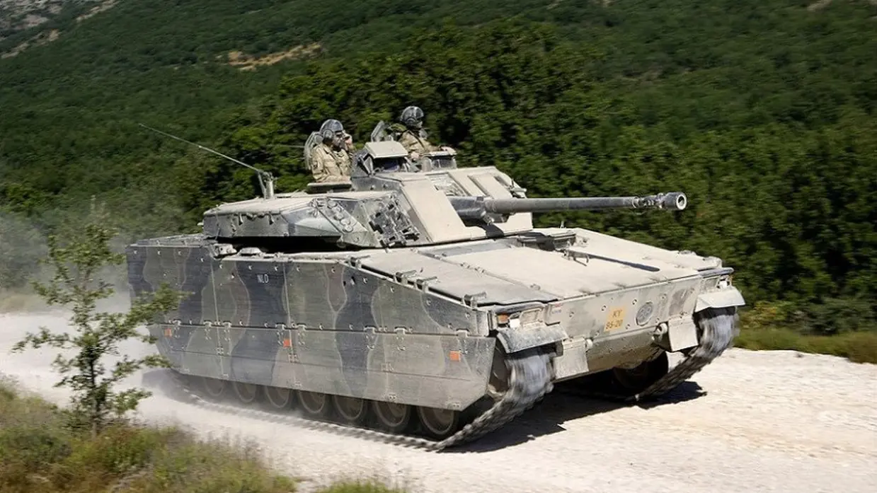  Een CV90-infanterievoertuig rijdt over de weg, een stofwolk achterlatend. Op de achtergrond zijn groene heuvels te zien.
