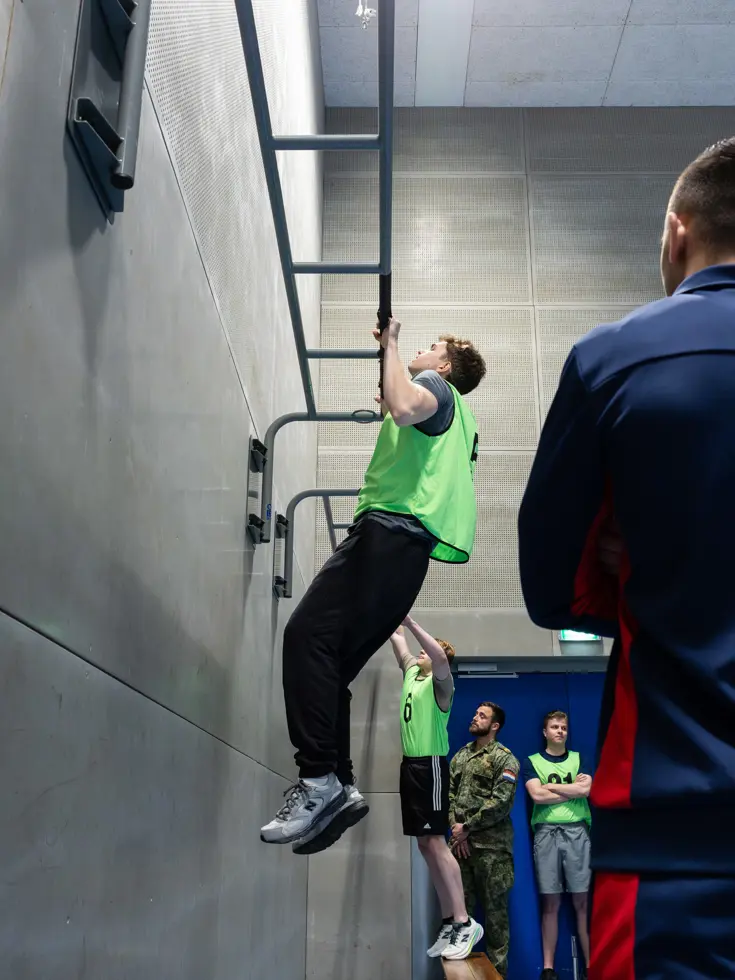 Jongeren in gele hesjes voeren diverse sportonderdelen uit in een sportzaal. 