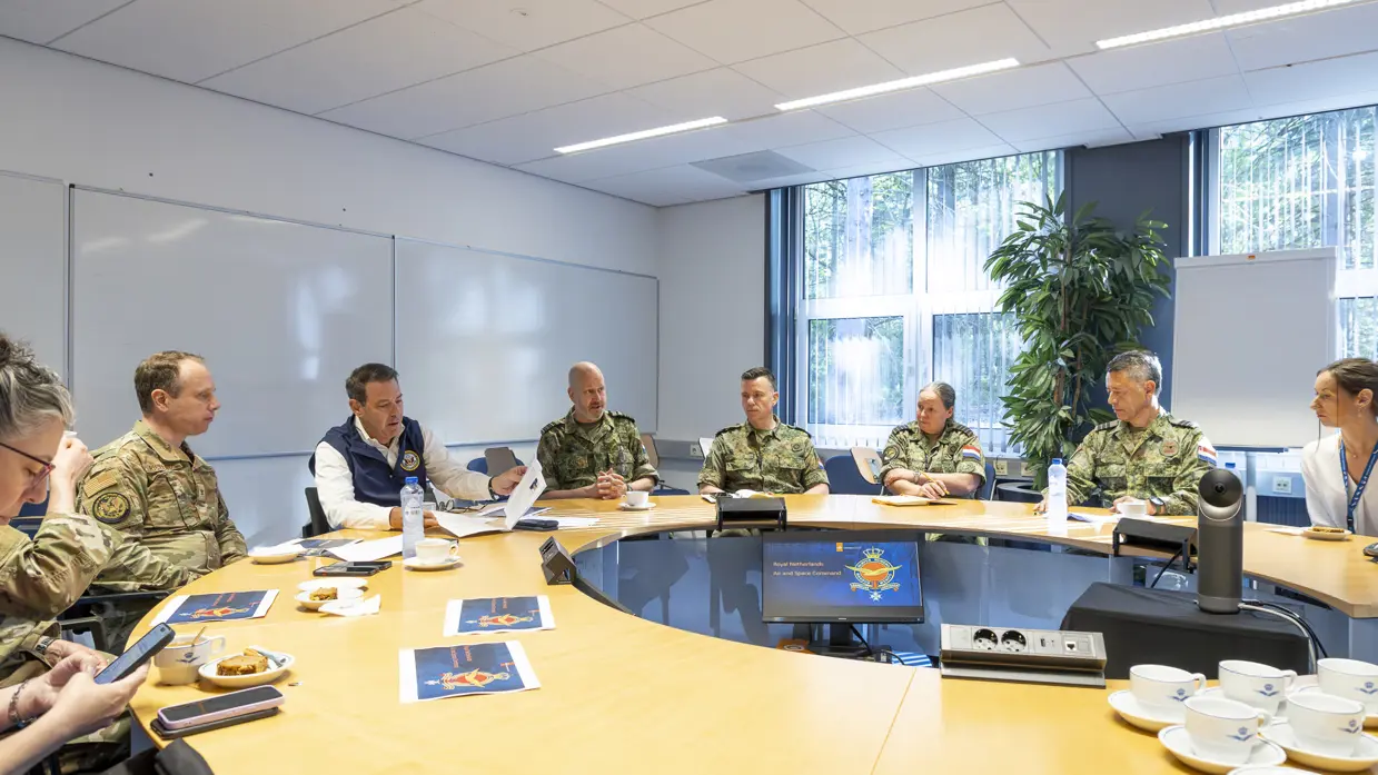 Popolo en Adang samen met nog een aantal militairen en een burger in gesprek aan een ronde tafel.
