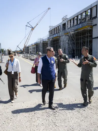 Popolo lopend in gesprek met een militair op een terrein met waarop gebouwen in aanbouw zijn. Achter hen loopt een aantal militairen en burgers met op de achtergrond een hijskraan.