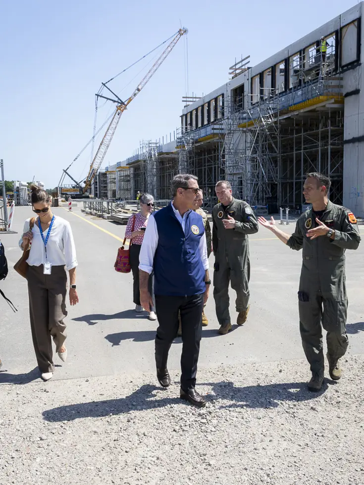 Popolo lopend in gesprek met een militair op een terrein met waarop gebouwen in aanbouw zijn. Achter hen loopt een aantal militairen en burgers met op de achtergrond een hijskraan.