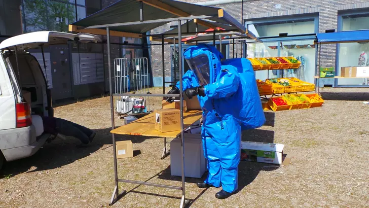 Persoon in beschermend blauw pak onderzoekt een pakketje in een marktkraam.