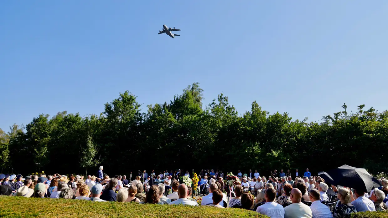 Publiek bij een plechtigheid kijkt omhoog naar een vliegtuig tegen de blauwe lucht op een mooie zonnige dag.