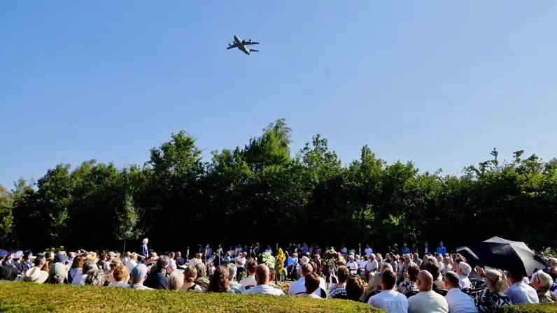 Publiek bij een plechtigheid kijkt omhoog naar een vliegtuig tegen de blauwe lucht op een mooie zonnige dag.