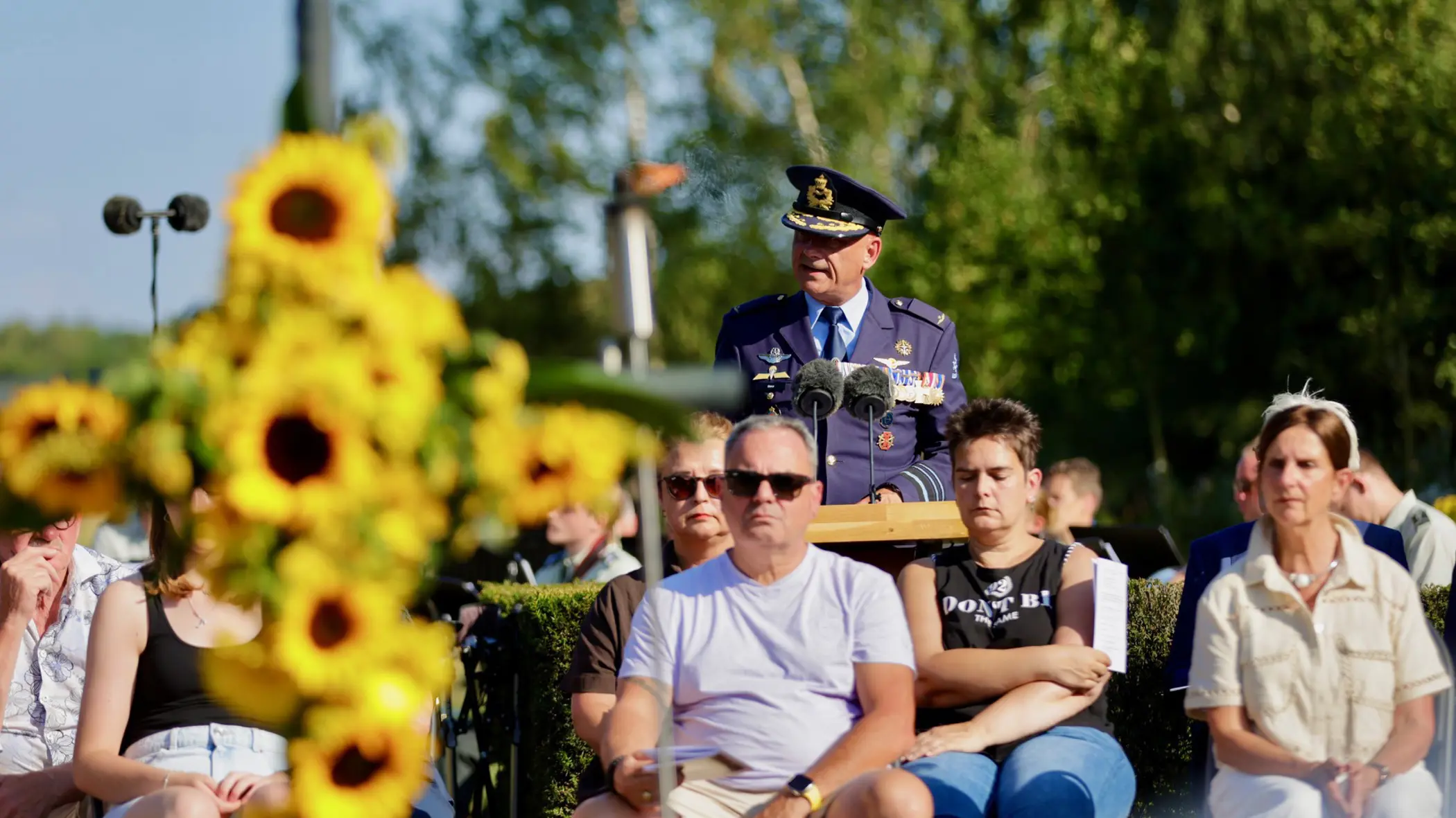 Een man in uniform achter een spreekgestoelte, met op de voorgrond mensen op stoelen en zonnebloemen op een zonnige dag.