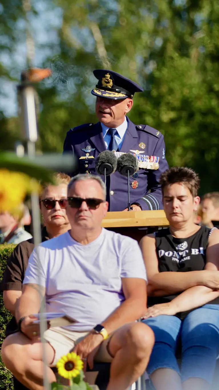 Een man in uniform achter een spreekgestoelte, met op de voorgrond mensen op stoelen en zonnebloemen op een zonnige dag.