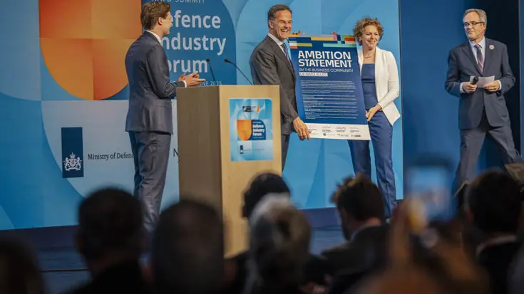 Man en vrouw houden kartonnen bord vast op podium. Links en rechts van hen kijken twee mannen toe.