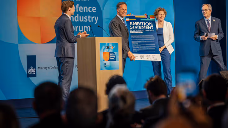 Man en vrouw houden kartonnen bord vast op podium. Links en rechts van hen kijken twee mannen toe.