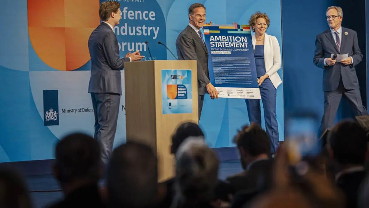 Man en vrouw houden kartonnen bord vast op podium. Links en rechts van hen kijken twee mannen toe.