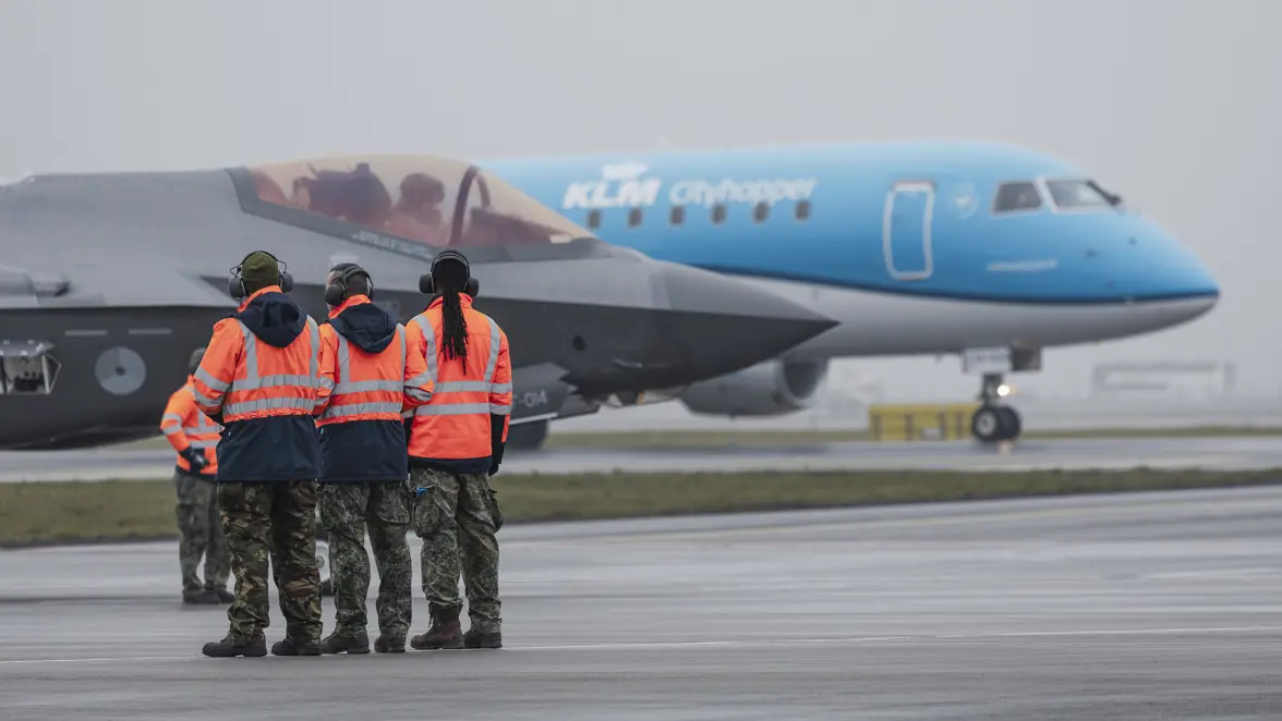 Vier mensen in een oranje veiligheidshesje kijken naar een geparkeerde F-35, waarachter een geparkeerd passagiersvliegtuig van KLM staat. 