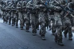 2 rijen marsende deelnemers aan de Nationale Weerbaarheidstraining in uniform met wapen in de hand.