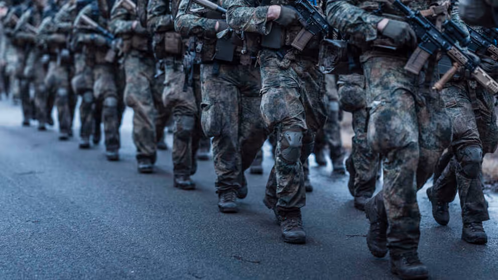 2 rijen marsende deelnemers aan de Nationale Weerbaarheidstraining in uniform met wapen in de hand.