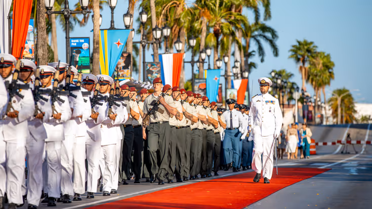 Een hoge militair loopt over een oranje loper in de zon en passeert tientallen militairen.
