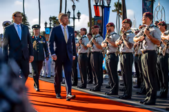 De koning loopt over een oranje loper in de zon en passeert een rij militairen met geweer.