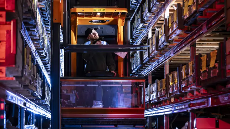 Een medewerker in een vorkheftruck kijkt naar de goederen in een hoogbouwmagazijn.