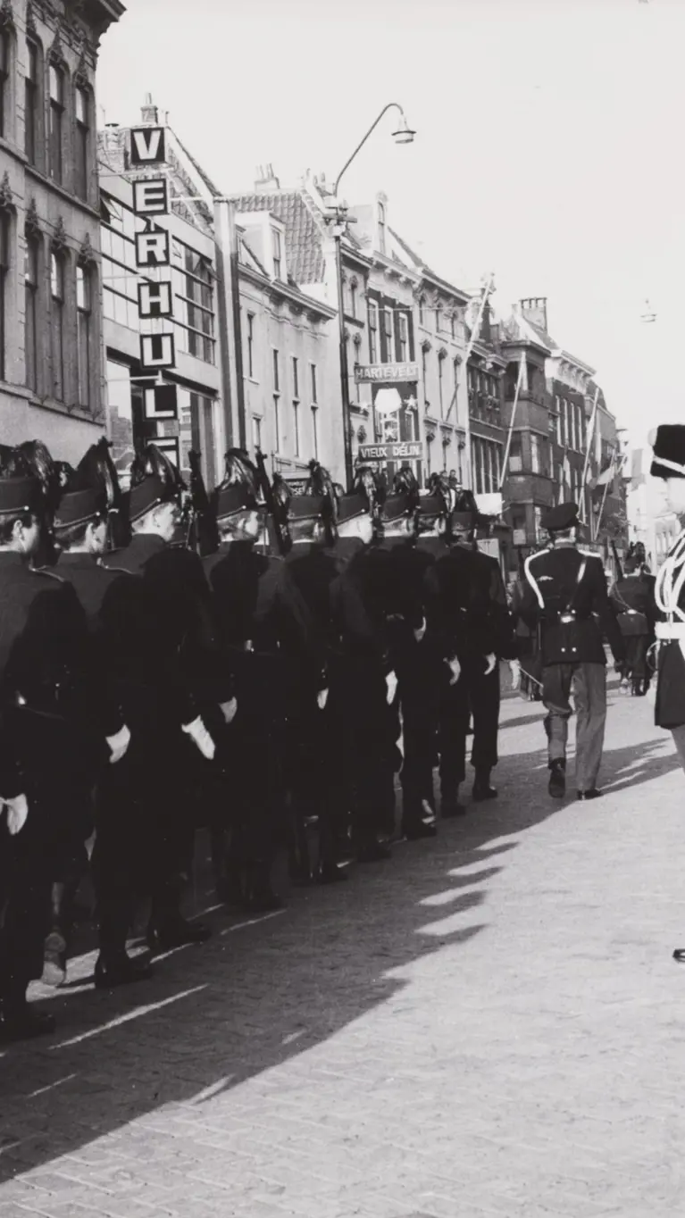 Een zwart-witfoto van een militaire parade met soldaten in uniform, marcherend door een straat met oude gebouwen en toeschouwers.
