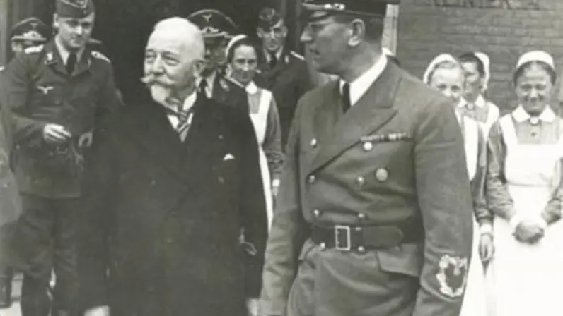 Een oudere man met snor en een man in militair uniform lopen samen, omringd door soldaten en verpleegsters bij een gebouw.